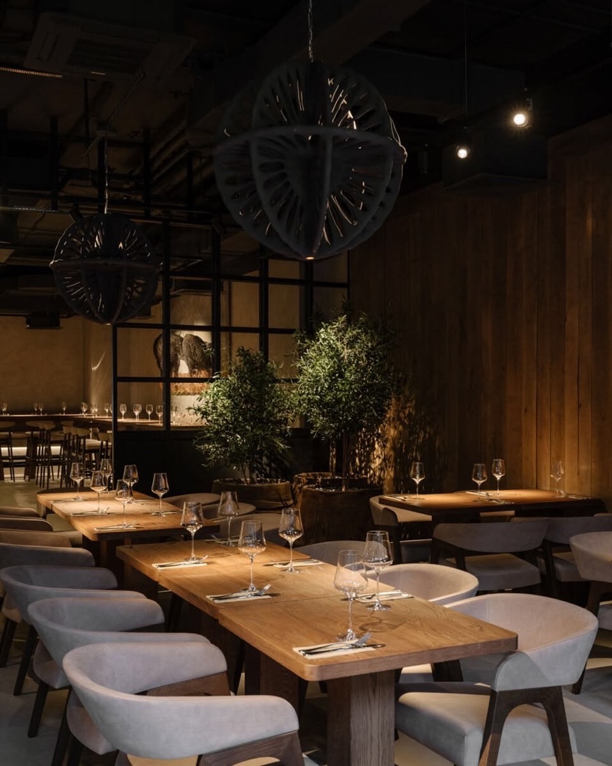 Salón de un restaurante con mesas de madera preparadas, copas y lámparas de gran formato.