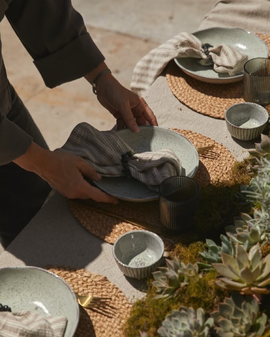 Dos manos colocan vajilla sobre una mesa, entre plantas y mantel de lino.